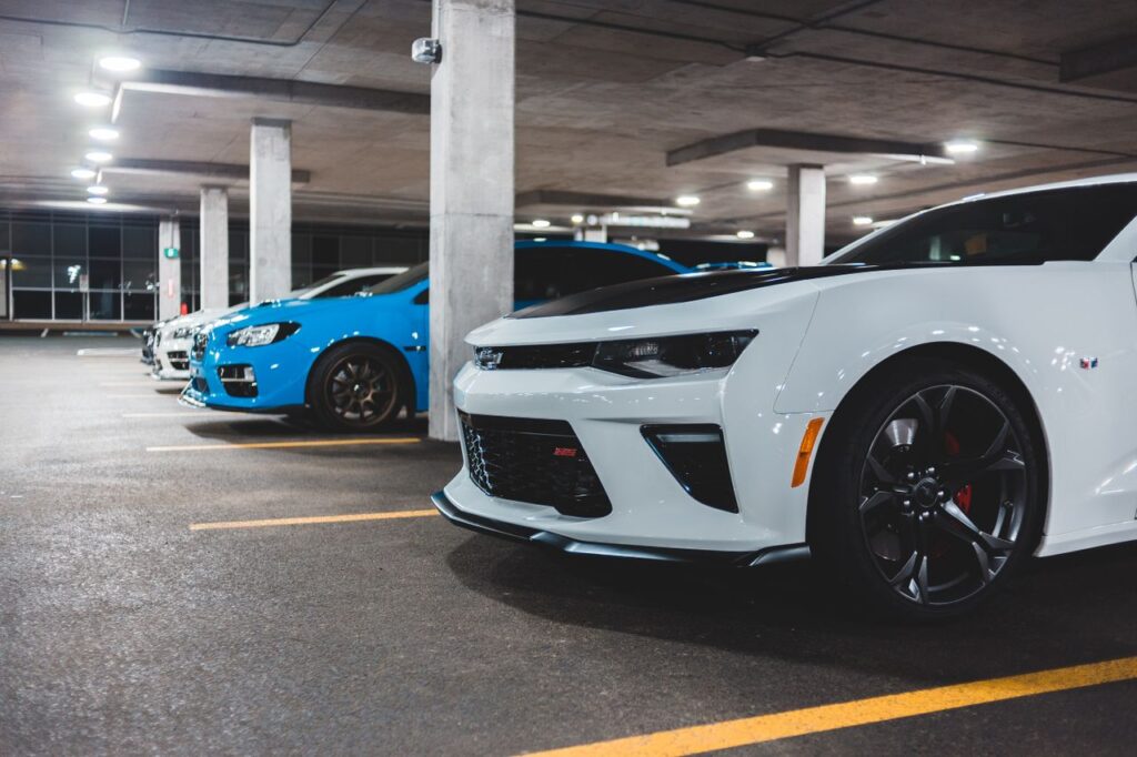 Front bumpers of contemporary luxury autos under roof with shiny lamps on asphalt parking in evening